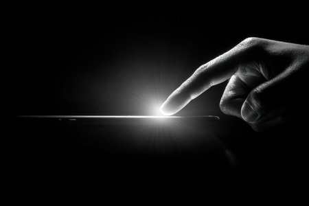 Artistic black-and-white photo depicting a hand touching a glowing smartphone. The strong light contrast emphasizes minimalist technology symbolism and modern digital interaction.の素材
