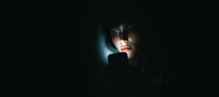A person holds a phone in a dark room, their face illuminated by a mobile banking app. The image captures themes of financial control, privacy, and modern solitude.の素材