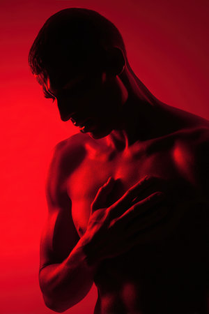A man holds his chest near the heart area, highlighted by dramatic red lighting. This cinematic photo realism image emphasizes health awareness and emotional intensity.の素材