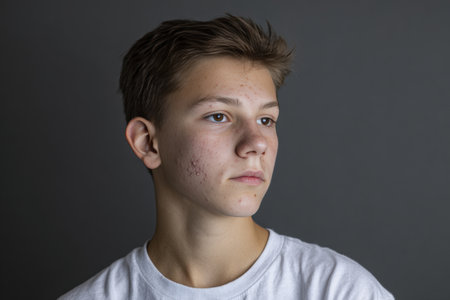 High-resolution studio portrait of a teen boy with acne scars and textured skin, wearing a plain white t-shirt. Set against a dark grey background, the image is deeply expressive.の素材