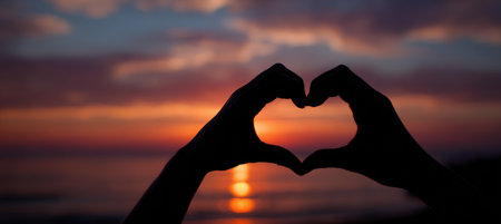 Silhouette of hands forming a heart shape against a vivid sunset. The horizon blends with warm colors, creating a poetic and romantic visual composition.の素材