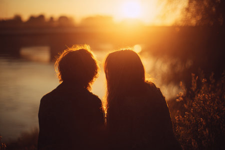A young couple sits by a river, enjoying a warm sunset. The backlight highlights their hair and silhouettes, creating a dreamy and emotional scene of romantic realism.の素材