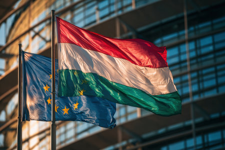 European Union and Hungary flags flutter under sunlight against a backdrop of mirrored windows, symbolizing diplomatic relations. The wide shot captures a stable, cinematic mood.の素材