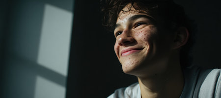 A teenage boy with acne-prone skin smiles warmly as daylight streams through a window, casting shadows that add depth to his features. He wears a simple t-shirt, exuding a natural charm.の素材