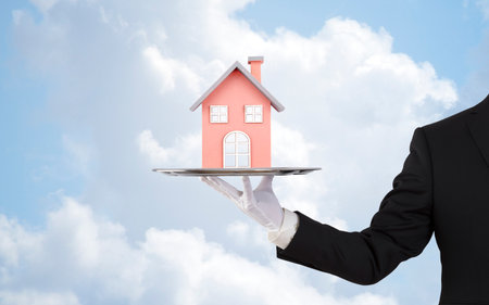 Businessman offering house model on silver tray over blue skyの写真素材