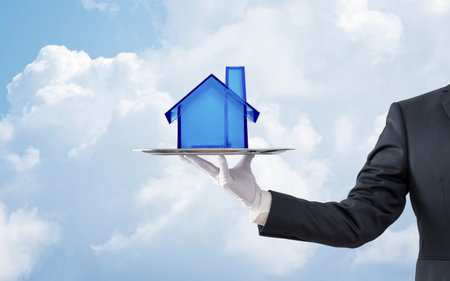 Businessman offering a blue crystal house model on a silver tray over blue skyの写真素材