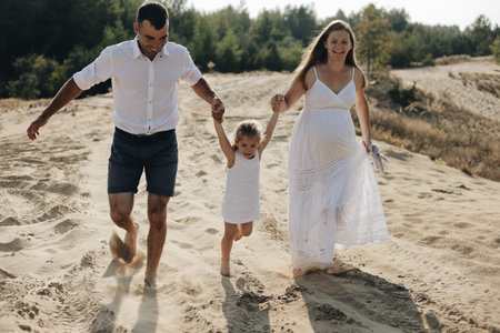 Cheerful caucasian family in white clothes with pretty daughter runs on desert sand dune. Pregnant motherの写真素材