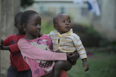 KENYA, KISUMU - MAY 20, 2017: Back view of little african girl carrying naked baby boy through the village.のeditorial素材