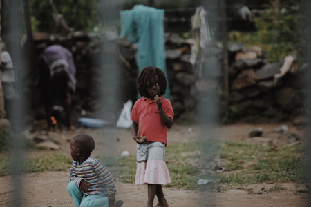 KENYA, KISUMU - MAY 20, 2017: View through the fence. Group of african people spending time outside. Little girl having fun.のeditorial素材