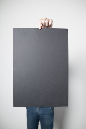 Man in a blue jeans is standing in front of the white wall while holding a black paper poster.
