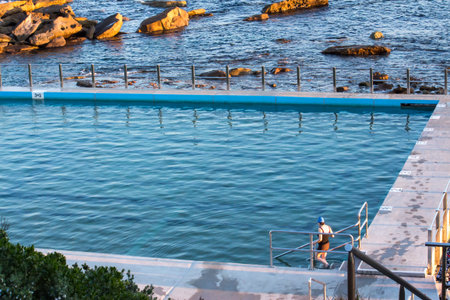 Woman entering seaside ocean swimming pool set against calm blue water and large exposed rocksの写真素材