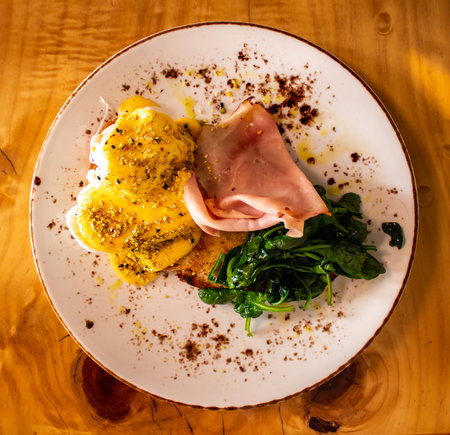 Breakfast meal plate of food comprising eggs Benedict with ham, spinach, toast and garnish set atop wooden tableの写真素材