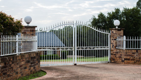 White metal wrought iron driveway property entrance gates set in brick fence, garden trees in...