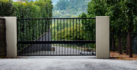 Black metal wrought iron driveway property entrance gates set in brick fence, concrete path, garden treesの写真素材