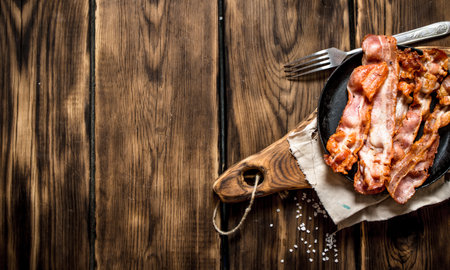 Fried bacon in a frying pan with a fork. On a wooden table.の写真素材