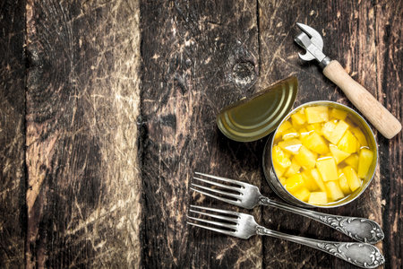 Canned pineapple in a tin can with forks and opener. On a wooden background.の写真素材
