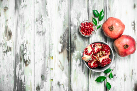 Grains and pieces of pomegranate in a bowl and whole pomegranates with leaves. On white wooden backgroundの写真素材