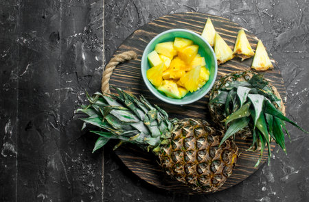 Slices of pineapple in a bowl and whole fresh pineapples on a tray. On black rustic backgroundの写真素材