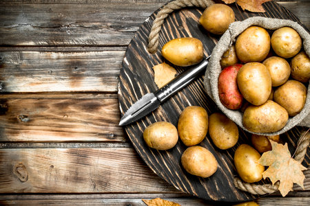 Potatoes in the sack on the tray with potato peeler. On wooden backgroundの写真素材