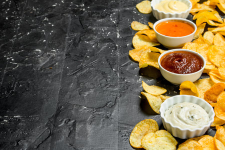 Potato chips with different sauces. On black rustic background.
