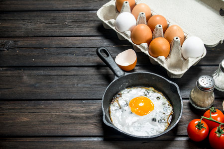 Fried egg in a pan with a cassette of fresh eggs. On a wooden background.の写真素材