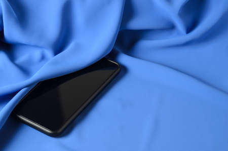 A black smartphone on a blue silk wavy background. The phone is on the bed on the blue sheet.