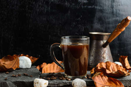 A glass mug of strongly brewed coffee on the stones with coffee beans and autumn leaves on the background of a coffee maker and dark wood. Selective focus.の写真素材