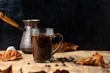 A glass mug of strongly brewed coffee on the stones with coffee beans and autumn leaves on the background of a coffee maker and dark wood. Selective focus.の写真素材