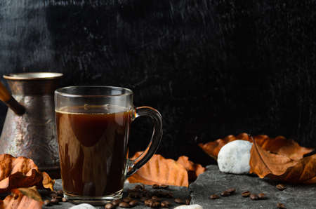 A glass mug of strongly brewed coffee on the stones with coffee beans and autumn leaves on the background of a coffee maker and dark wood. Selective focus.の写真素材