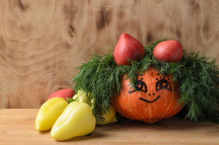 Funny pumpkin with painted eyes and a smile on a wooden background in the decoration of dill and vegetables. Selective focus.の写真素材