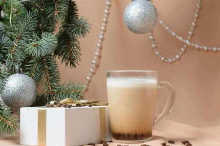 Christmas. A New Year's composition made of branches of a Christmas tree, decorated with silver balls, a gift box, a glass mug with coffee and milk on the background of flesh-colored waves. Selective focus.の写真素材