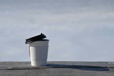 An abandoned tea paper cup with a black lid on a wooden bench with a blurred snow background. Selective focus.の写真素材