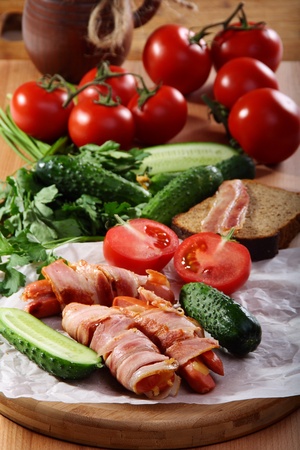 Sausages wrapped in bacon with vegetables and herbs on a wooden table.の写真素材