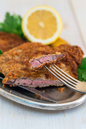 Piece of beef schnitzel on a fork, parsley and lemon on metal tray.