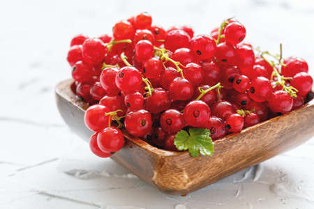 Red currant in a wooden bowl on a white concrete background.