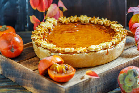 Homemade pumpkin pie, juicy persimmons and colorful autumn leaves on dark wooden background.