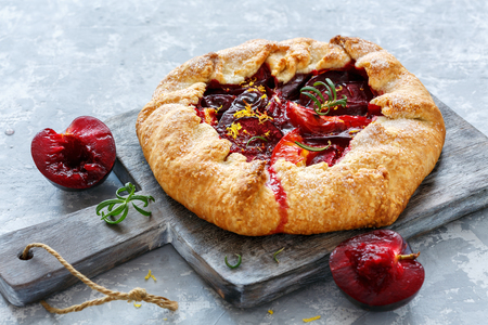 Free-form pies (Galette) with yellow and red plums, rosemary, lemon zest on a grey concrete background.の写真素材