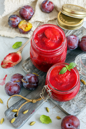 Glass jars of homemade plum jam with cardamom and plums in an old wooden bowl on textured concrete background.の写真素材