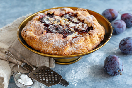 Bronze pie stand with plum filling and almond flakes and fresh blue plums on textured concrete table.の写真素材