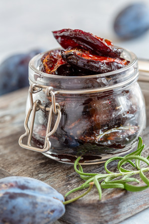 Spicy dried plum in jar and sprig of rosemary on a wooden serving board, selective focus.の写真素材
