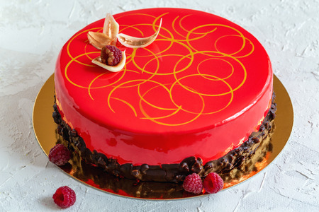 Mousse cake with red mirror glaze, chocolate decor and fresh raspberries on a textured white background.の写真素材