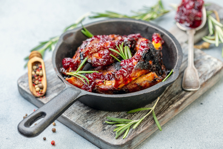 Cast iron pan with baked pork ribs, rosemary, spoon with cherry sauce on wooden serving board, selective focus.の写真素材