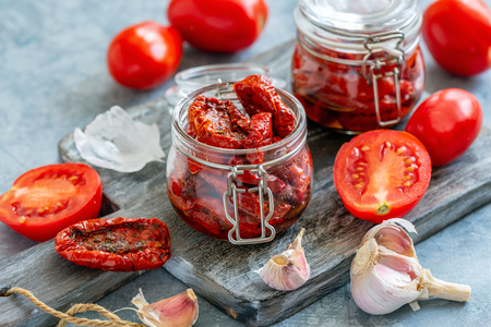 Jars of spicy sun-dried tomatoes, fresh tomatoes, garlic on a wooden chopping board, selective focus.の写真素材