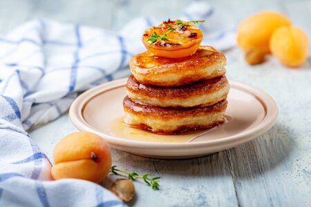 Thick pancakes or fritters are served with apricot jam and caramelized apricot on a ceramic plate, selective focus. White wooden background.の写真素材