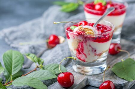 Glasses with cherry cheesecake on an old wooden serving board and dessert spoons on a textured gray background, selective focus.の写真素材