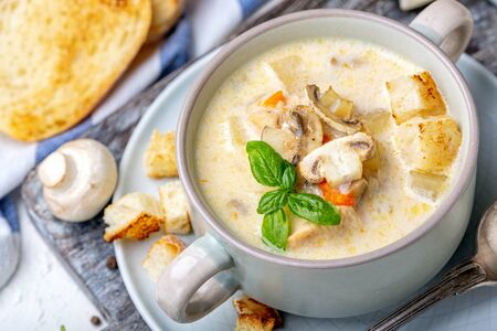Homemade chicken cream soup with mushrooms, cheese, croutons and green basil in a ceramic saucepan on a wooden serving board, selective focus.の写真素材
