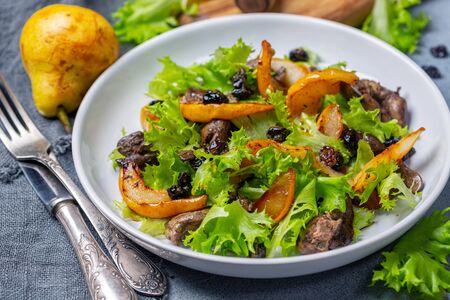 Warm salad with crispy lettuce leaves, fried chicken liver, caramelized pear slices, raisins and balsamic sauce in a white plate close-up, selective focus.の写真素材
