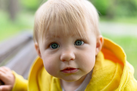 Close-up of a young child with striking blue eyes and blond hair, wearing a bright yellow jacket,...