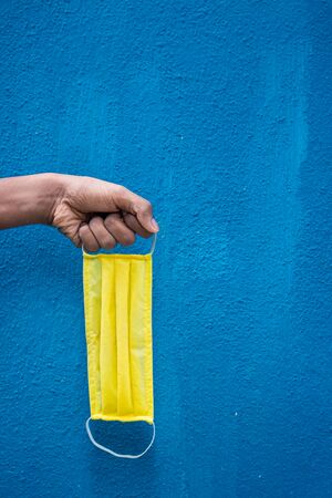 Man holding yellow face mask holding on his hand isolated on Blue background. Protective face mask on hand against Covid-19. Color face mask.Space for textの写真素材