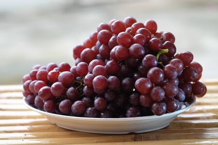 Large purple grapes are placed in a white plate on a bamboo table.の写真素材